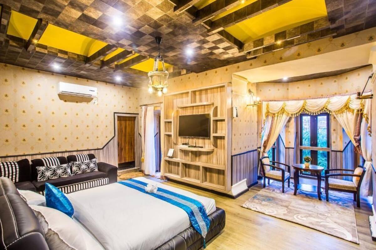 Habitación de hotel con una cama grande, televisión, mueble de madera para entretenimiento, sofá y balcón con cortinas.