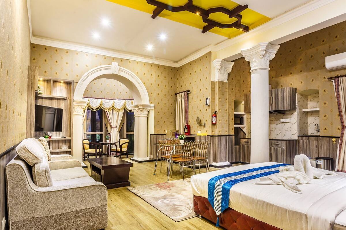 Habitación de hotel con cama, sofá, mesa de comedor con sillas, televisor y área de cocina.