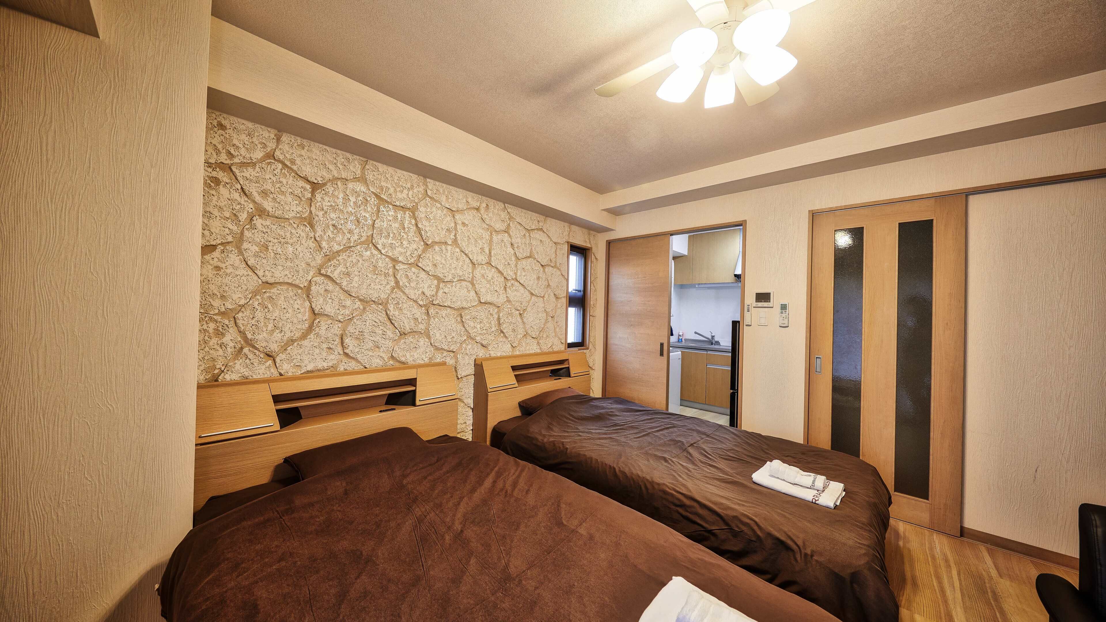 Une chambre d’hôtel avec deux lits, un mur en pierre, un ventilateur de plafond et une petite kitchenette.