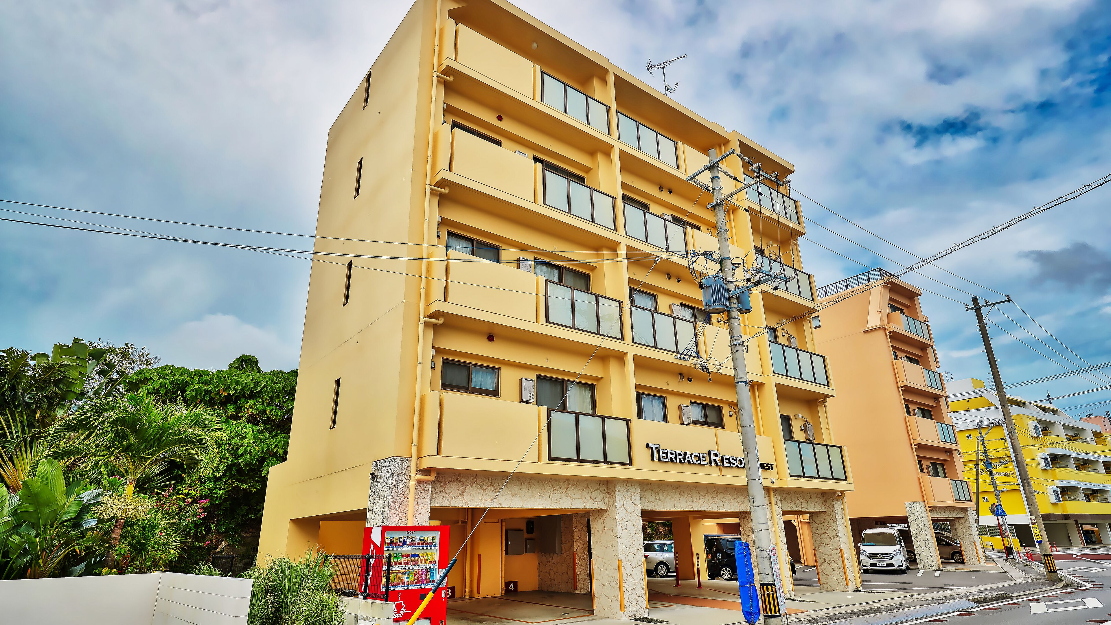 Un hotel a più piani con facciata gialla, numerosi balconi e una macchina distributrice all'esterno.