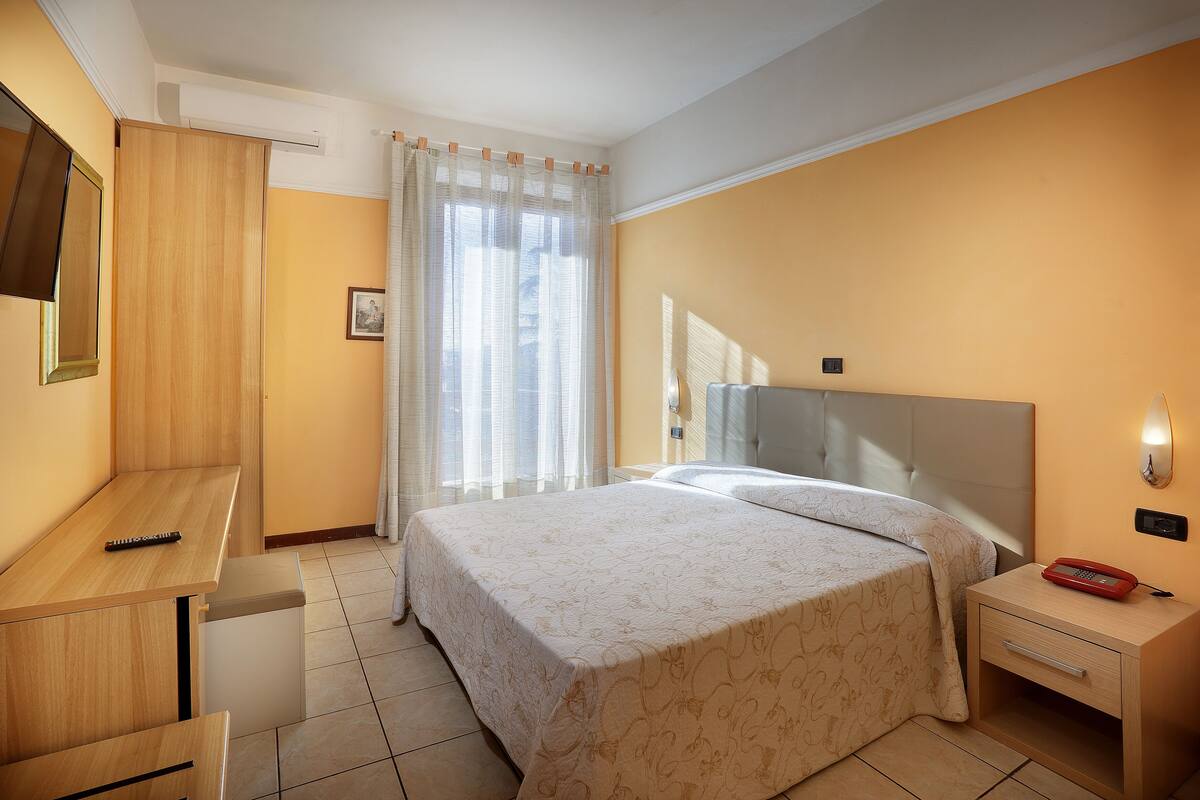 A hotel room with a bed, a desk, a chair, a nightstand, and a bedside table.