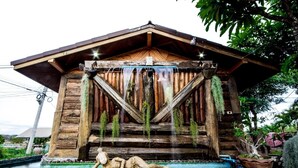 Ein rustikales Holzgebäude mit einem geneigten Dach, einem Wasserspiel mit hängenden Pflanzen und einer Steinskulptur im Eingangsbereich.