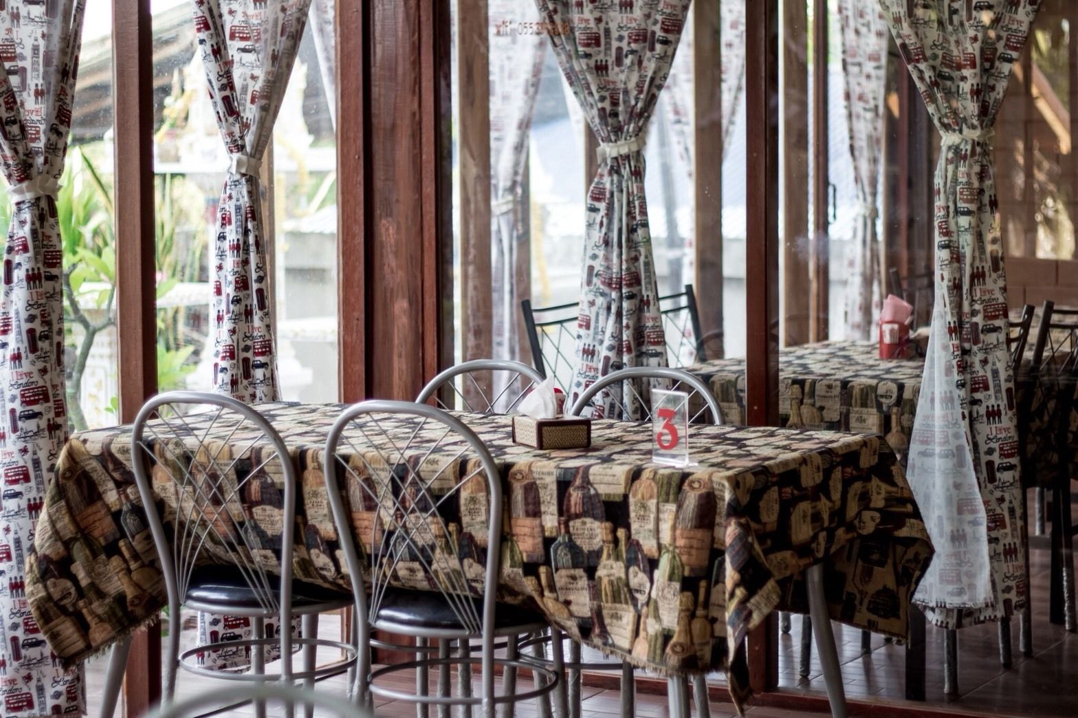 Un restaurant où une table est recouverte d’une nappe imprimée avec un motif de vieux journal, avec des chaises en métal et un panneau portant le chiffre 3 posé sur la table.