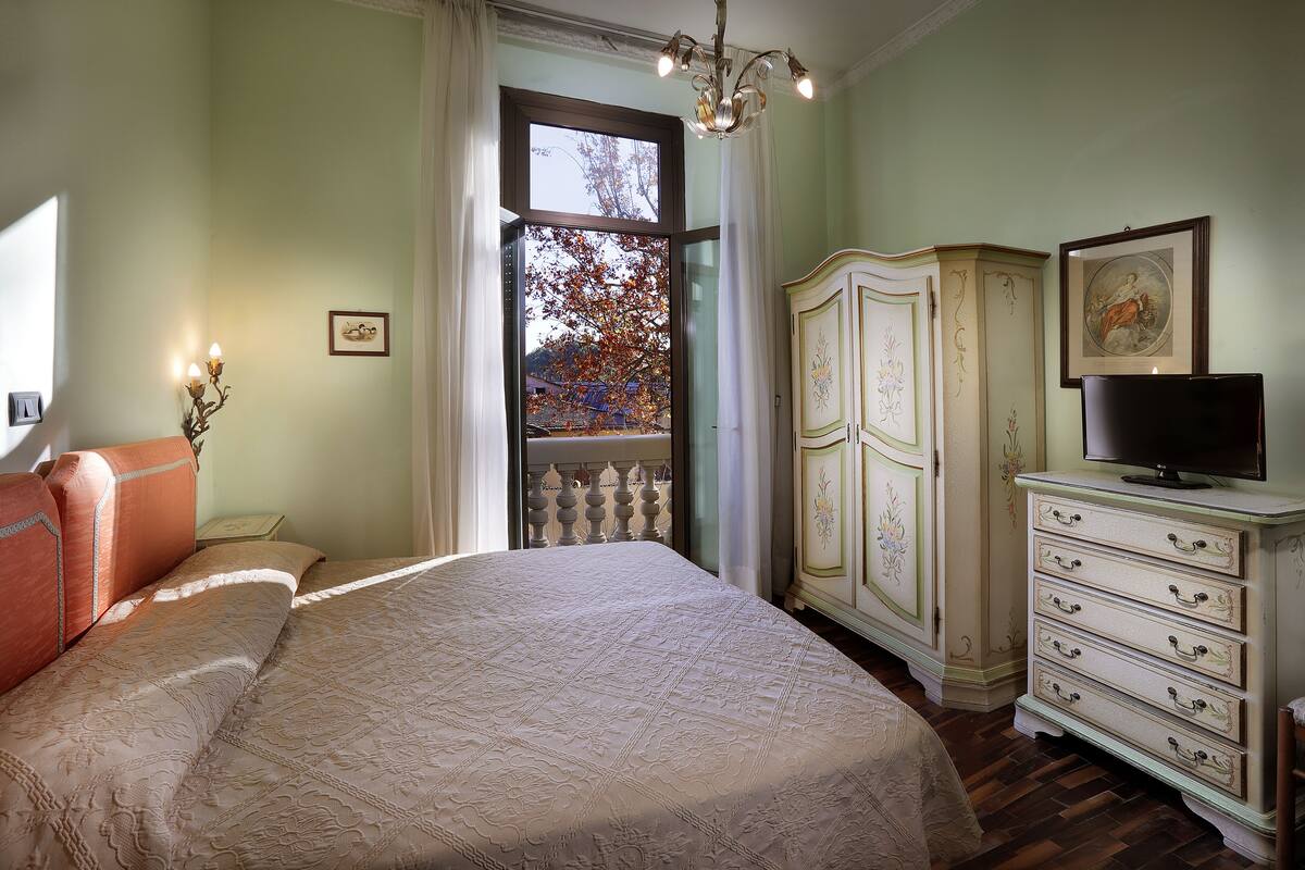 A bedroom with a large bed, a dresser with drawers, a television on top, and a window with a view of trees and a balcony.