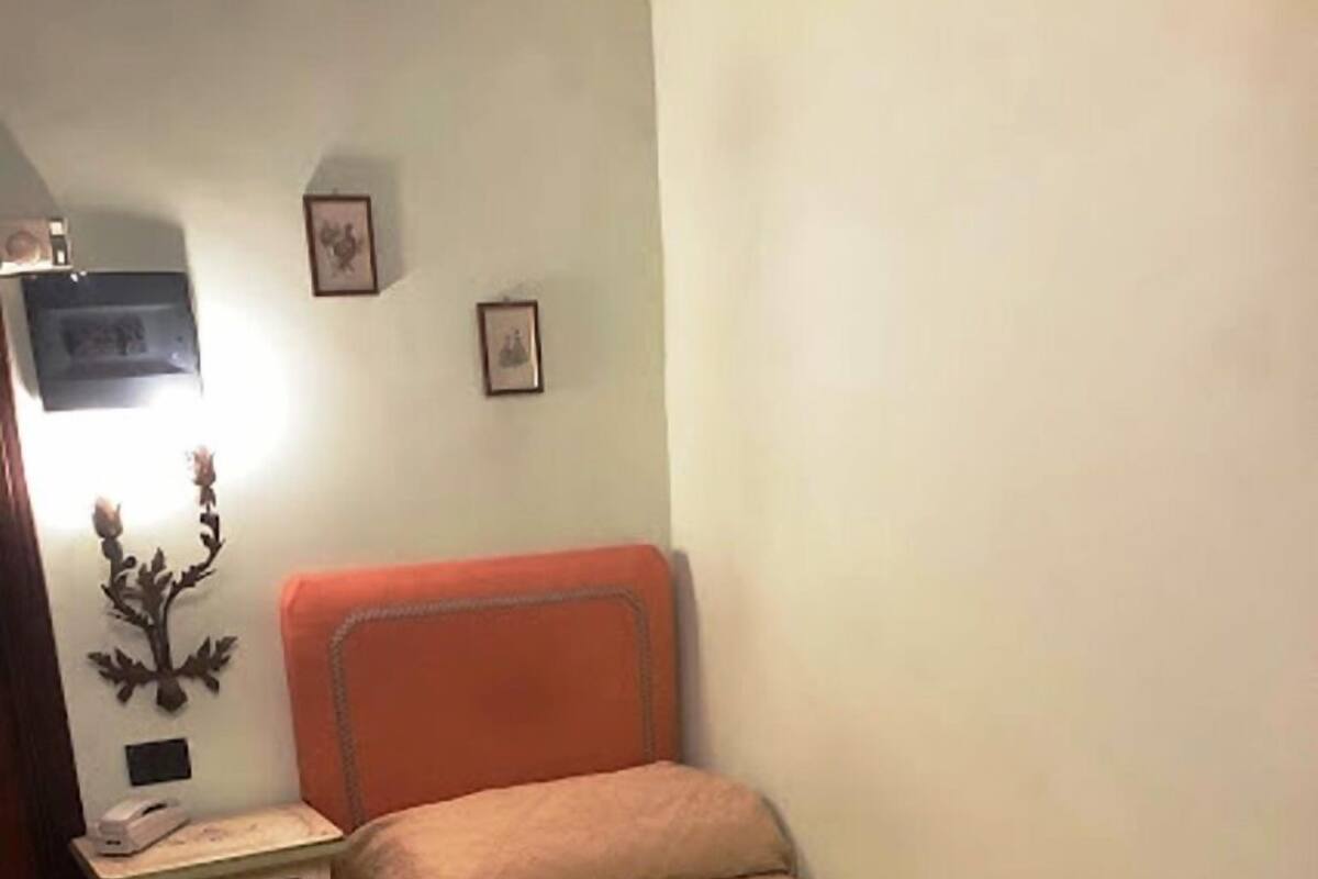 A small, single-bed room with a wooden floor, a nightstand with drawers, a lamp, and two framed pictures on the wall.