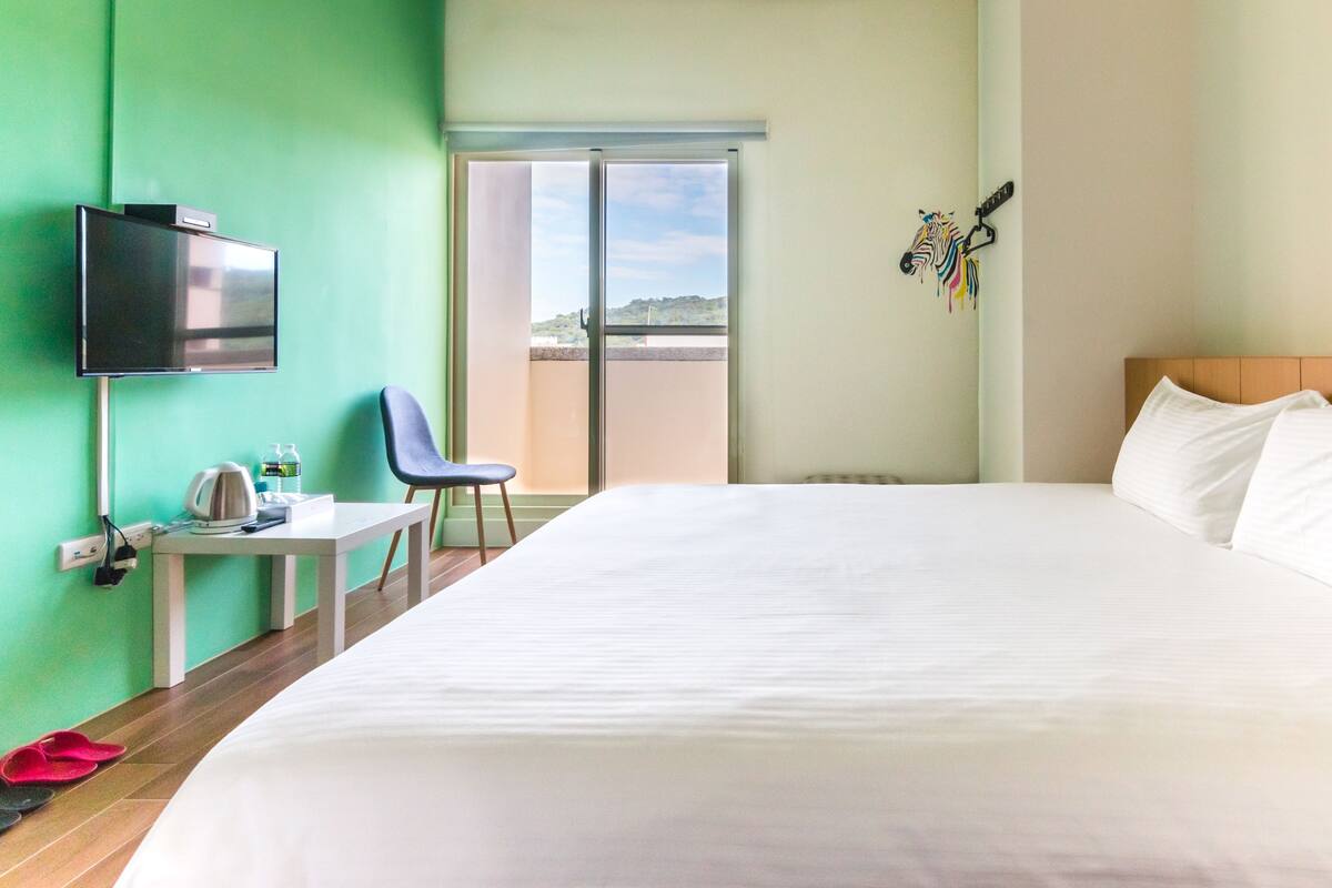 A hotel room with a large bed, a desk with a chair, a television, and a sliding door to a balcony.