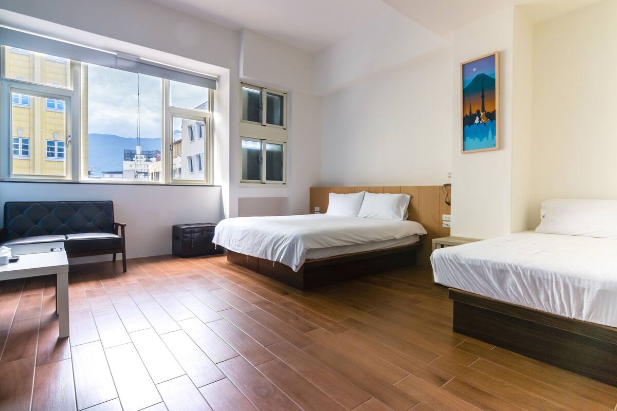 A hotel room with two beds, a sofa, a table, and large windows offering a view of buildings and mountains.