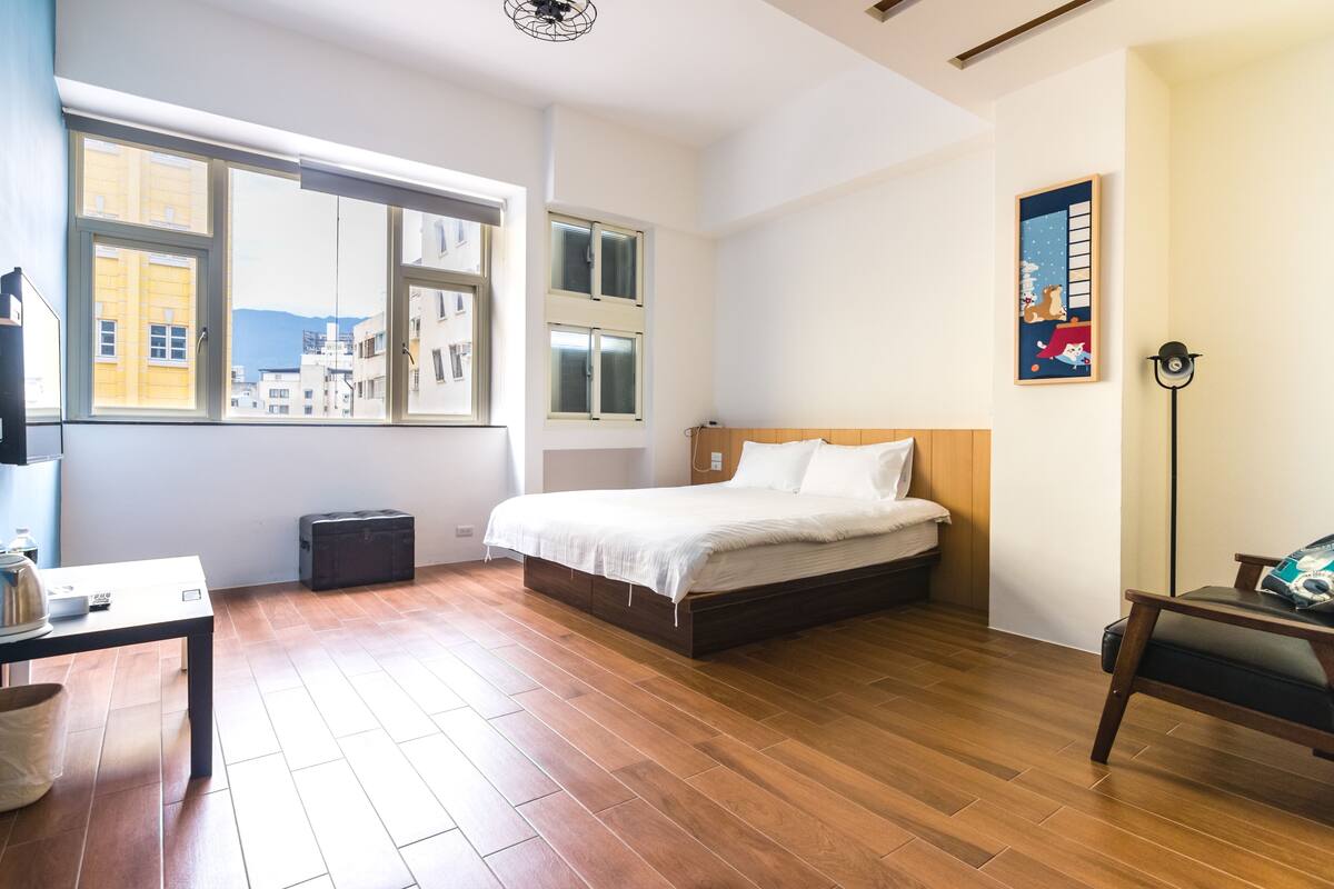 A bedroom with a wooden floor, a bed with white bedding, a desk with a kettle, and a suitcase.