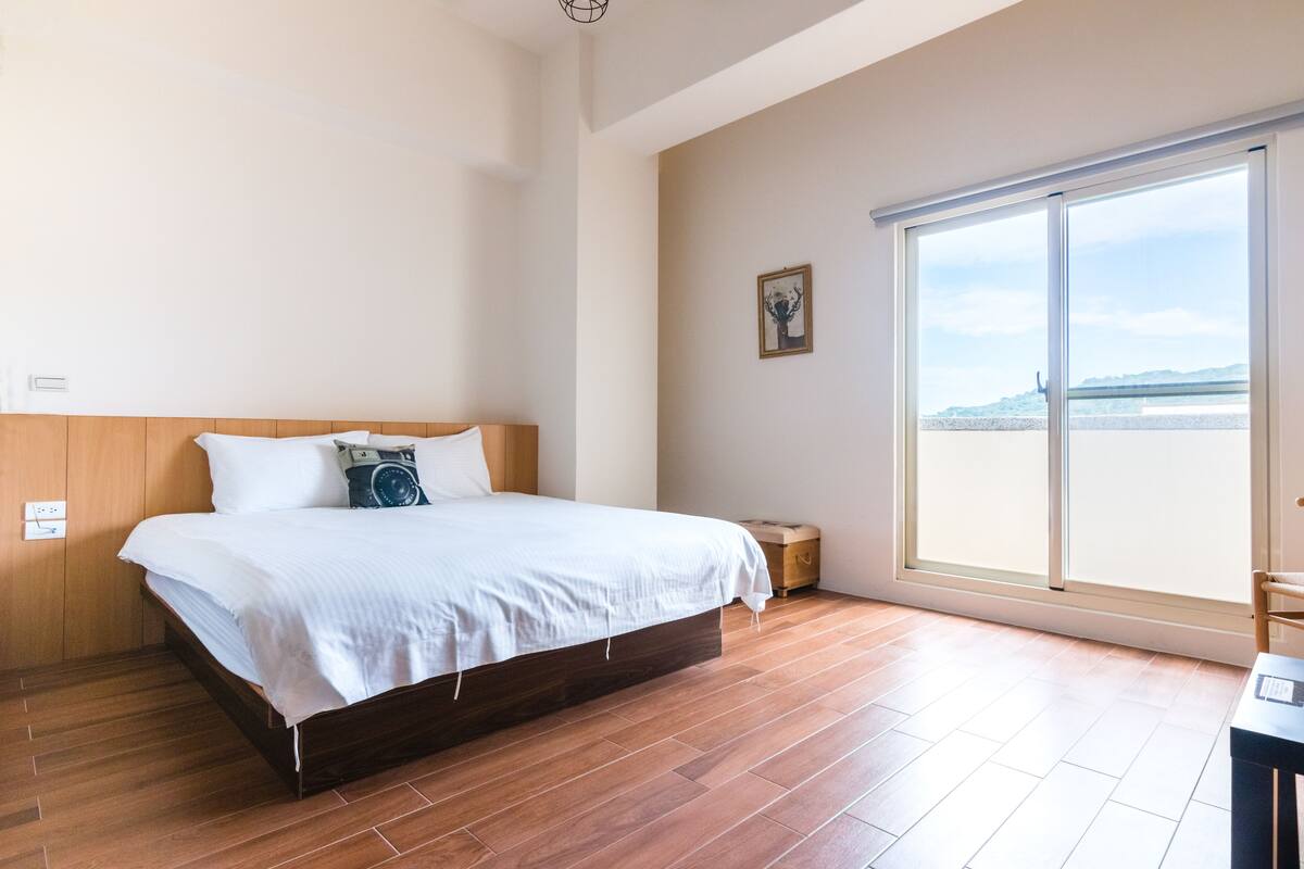 A bedroom with a large bed, wooden flooring, a window with a view, and a small table.