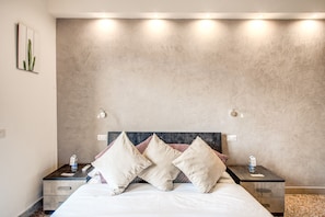 A neatly made bed with three pillows, a bedside table with a water bottle, and a wall-mounted light fixture.