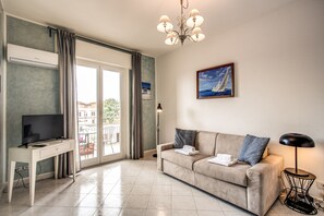 Un salon comprenant un canapé, un téléviseur, un bureau, un lustre, un tableau et un balcon avec des rideaux.