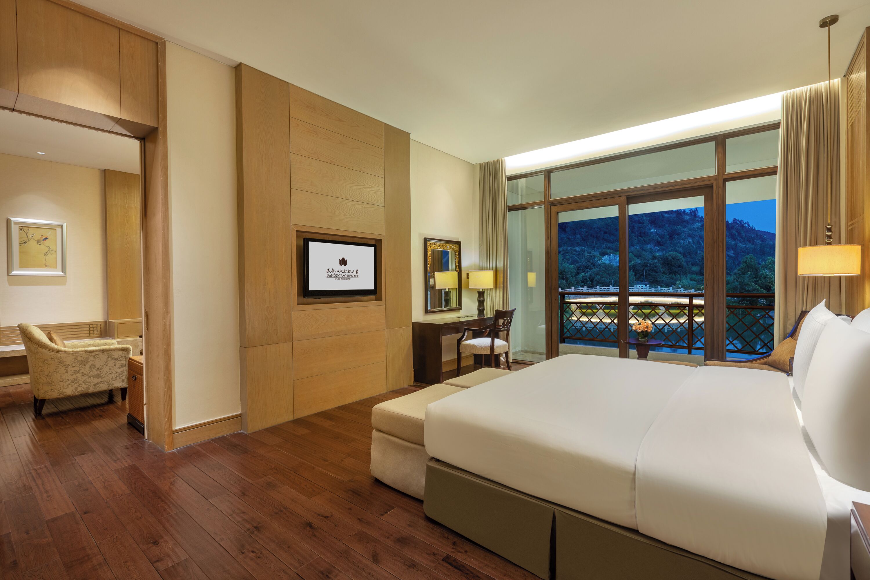 A hotel room with a large bed, a desk with a chair, a television, and a balcony with a view.