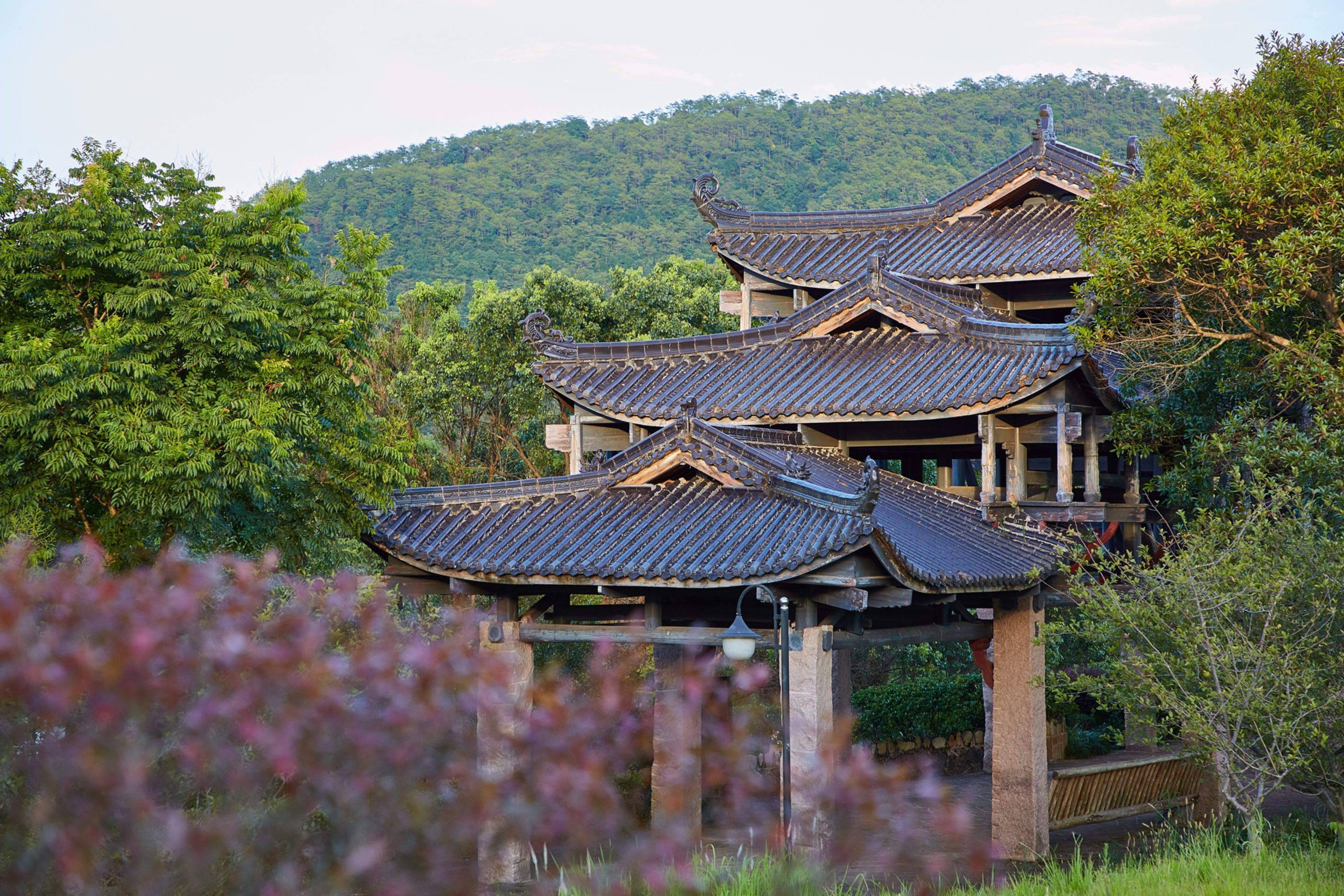 A traditional multi-tiered building with a tiled roof, surrounded by lush greenery and hills.