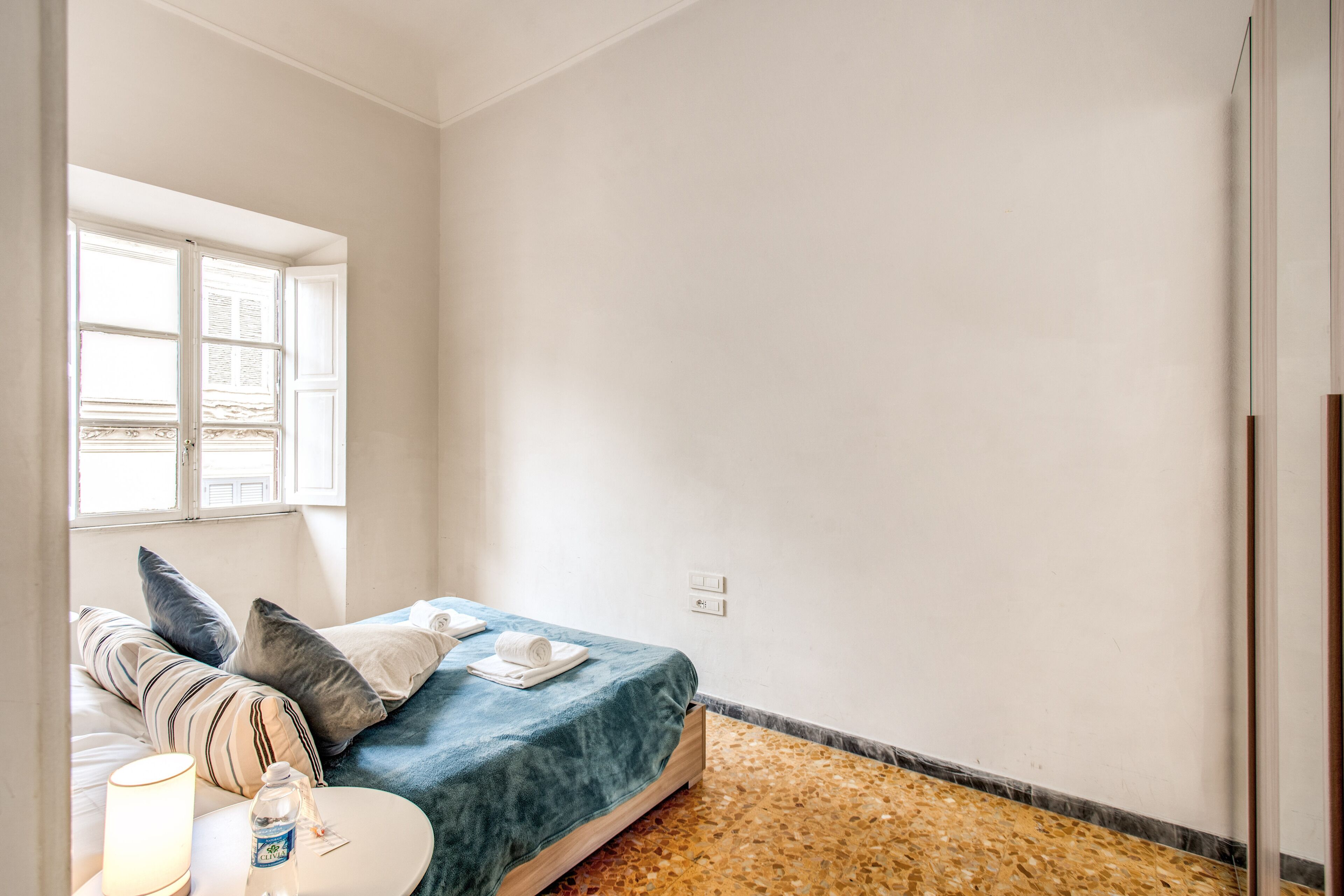 A bedroom with a bed, pillows, a bedside table with a lamp, and a window.