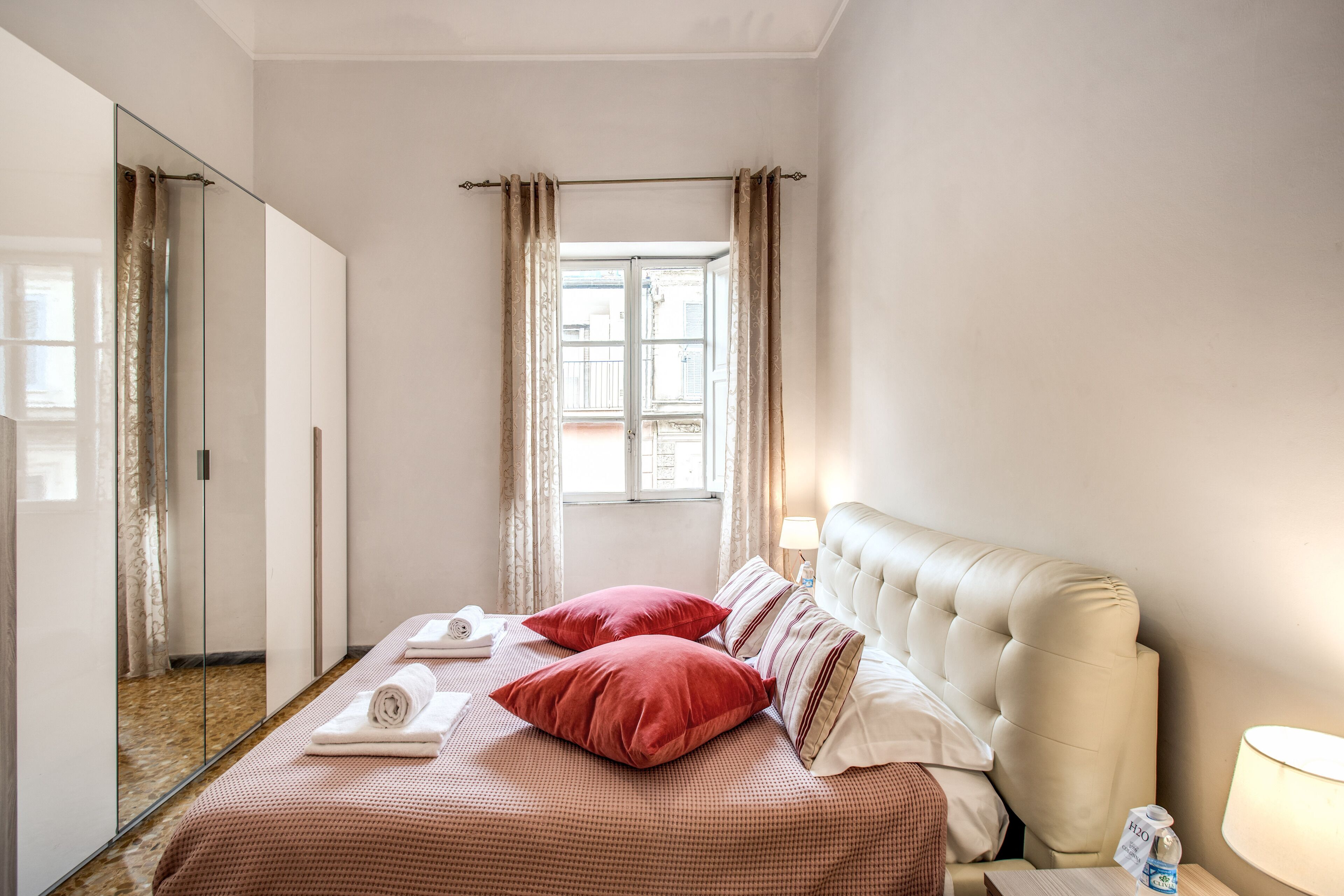 A hotel room with a bed, a bedside table with a lamp, a window with curtains, and a mirror.