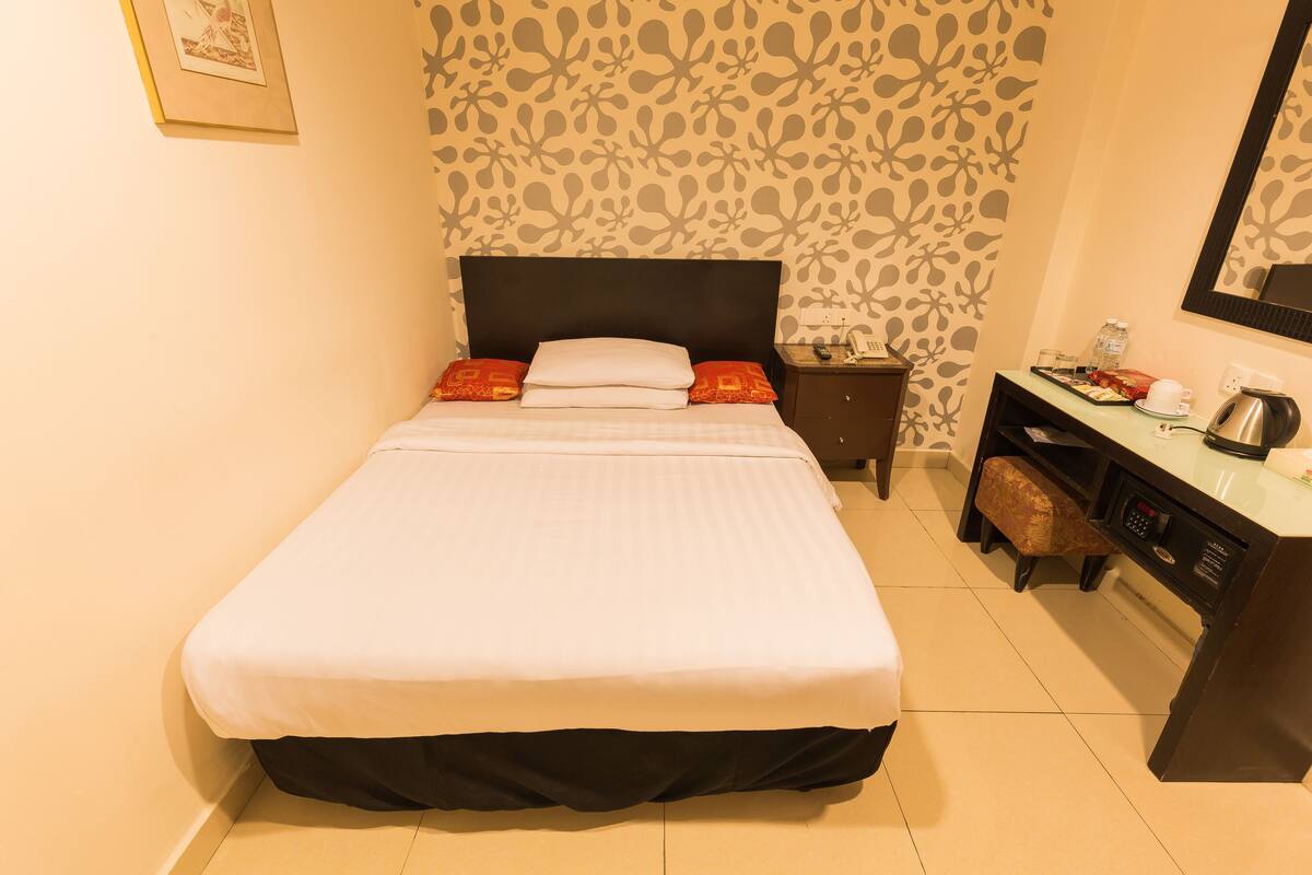 A neatly made bed with a black headboard, a nightstand with a lamp, a small table with a cup and a kettle, and a patterned wallpaper.