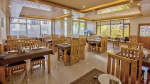 A spacious dining area with wooden tables and chairs, large windows offering natural light, and a modern ceiling design. - Hotel Wild Rose (Manali)