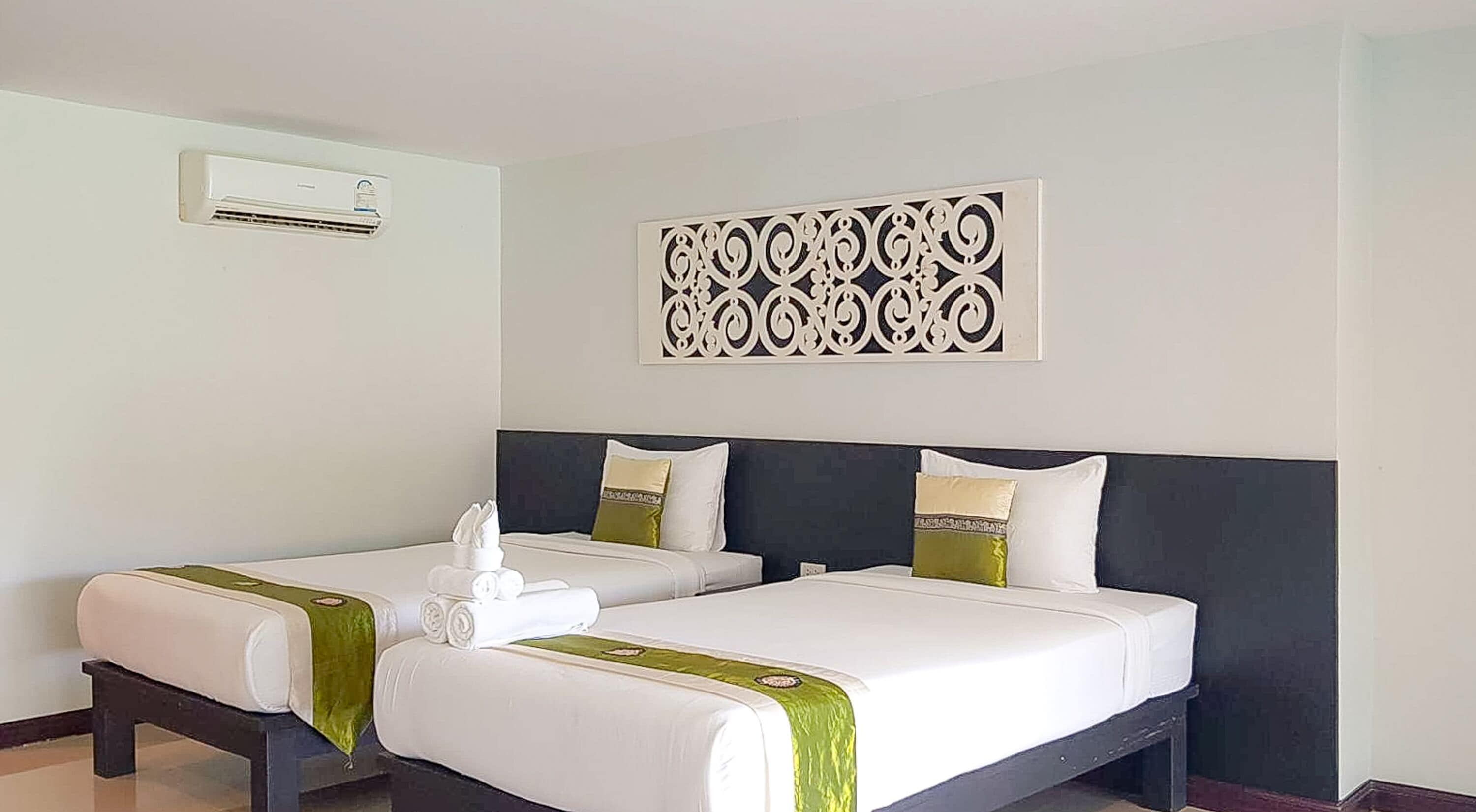 Habitación de hotel con dos camas, un adorno de pared, un equipo de aire acondicionado y una cama bien tendida con una toalla.