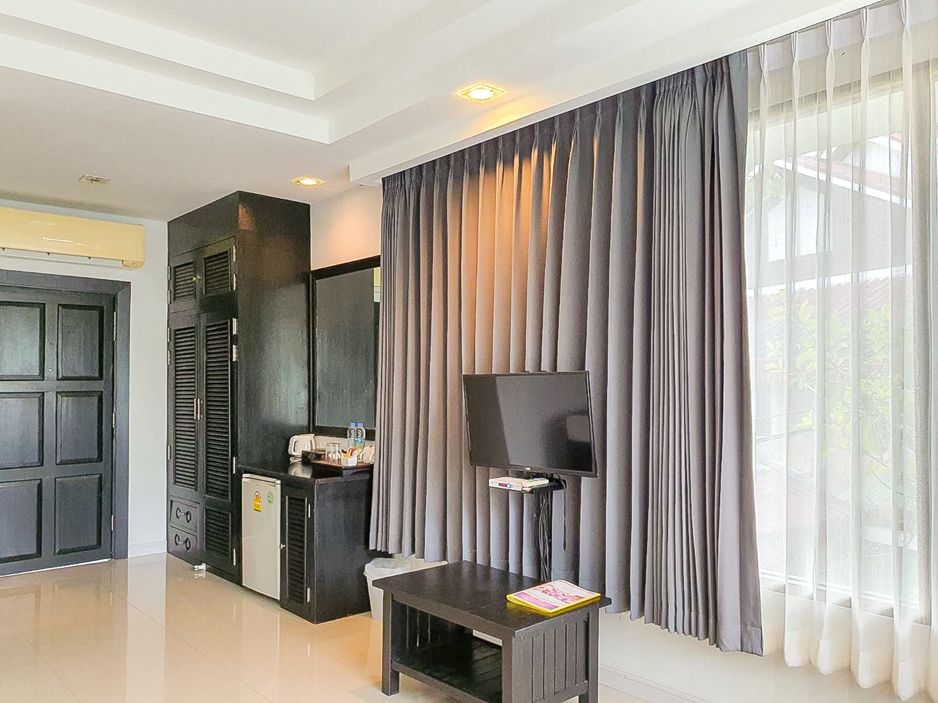 A modern hotel room with a large window, a flat-screen TV, a small table, and a minibar.
