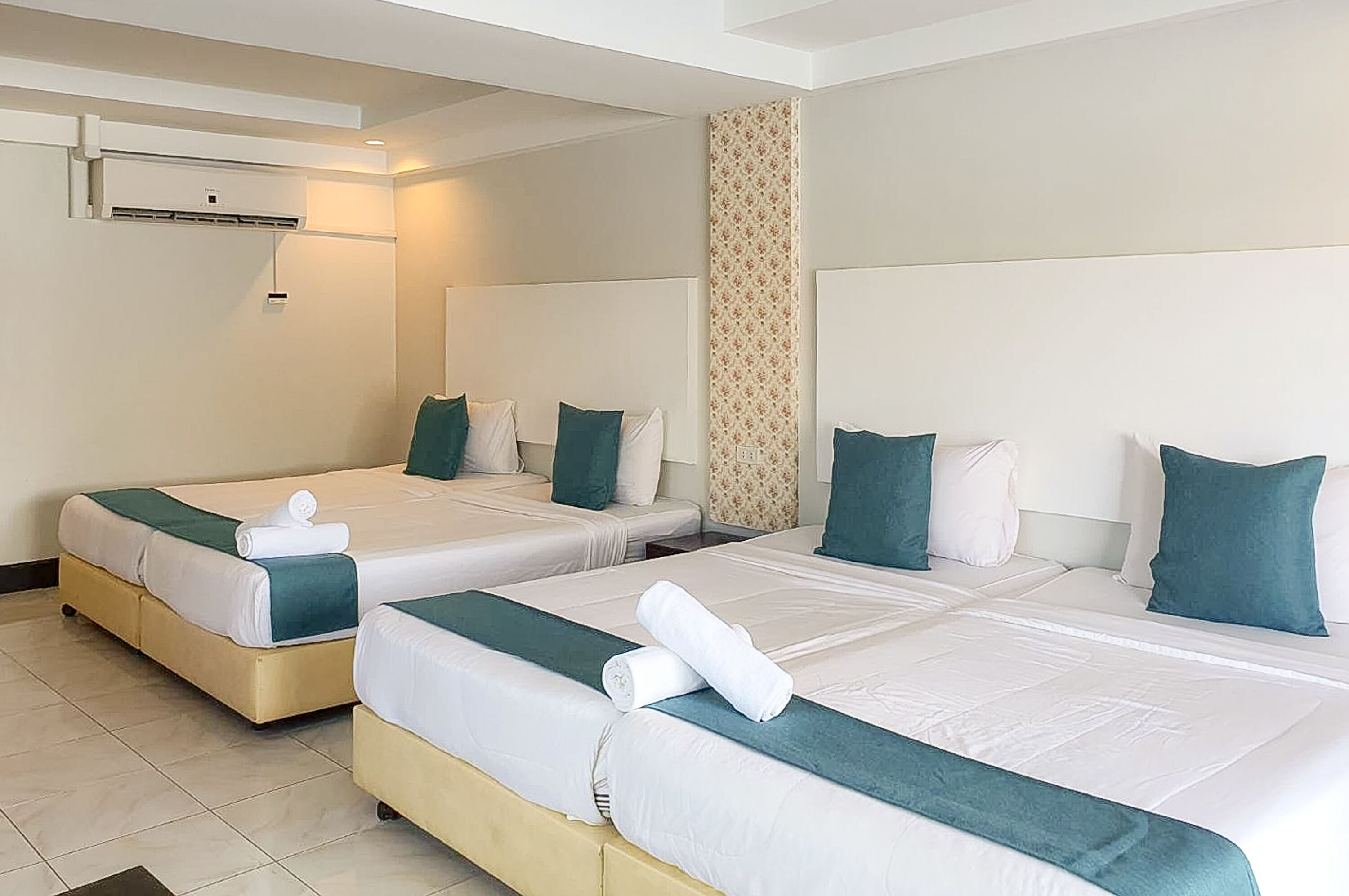 Habitación de hotel con dos camas, cada una con ropa blanca y detalles azul verdosos, aire acondicionado de pared y un columnario de papel tapiz estampado.