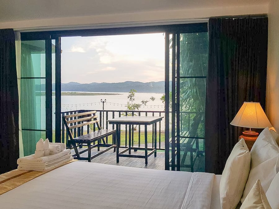 A hotel room with a bed, a table, and chairs, a balcony with a view of a lake and mountains.