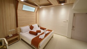 A hotel room with a bed, a nightstand, a desk, a chair, a ceiling fan, and a door. - Hotel Palmyra Grand Inn (Tirunelveli)