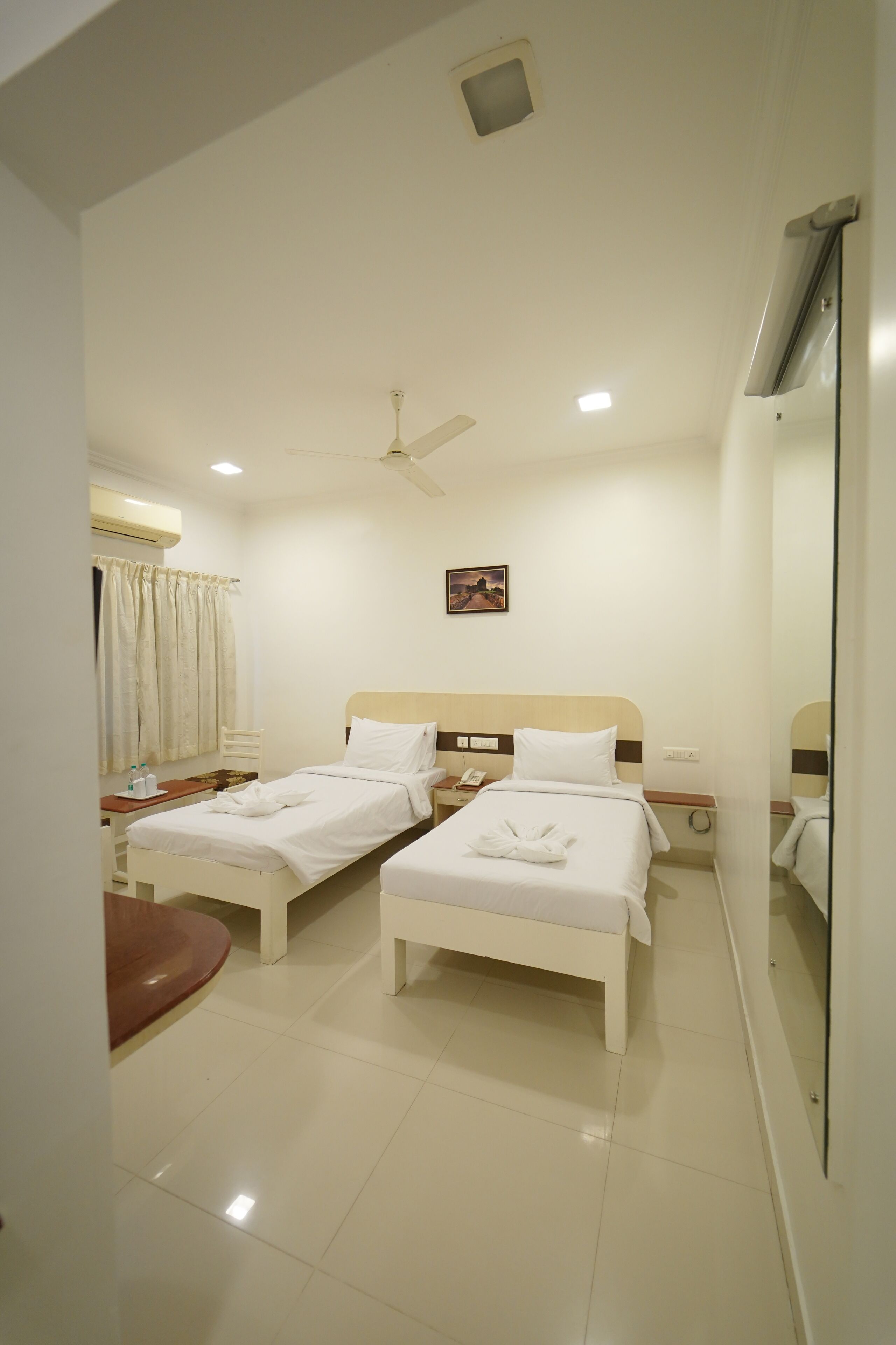 A hotel room with two beds, a bedside table, a mirror, a ceiling fan, and a window with curtains.