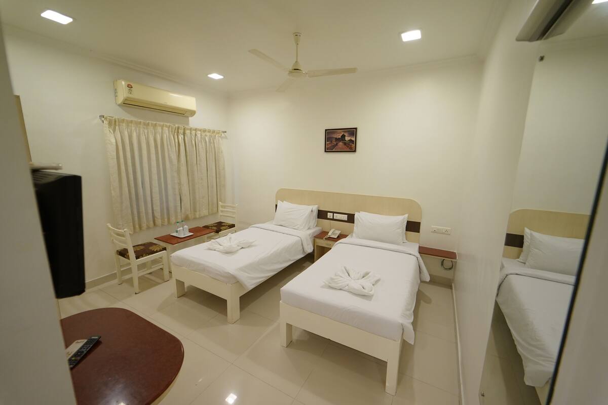 A hotel room with two single beds, a desk, a chair, an air conditioner, a ceiling fan, and a small painting on the wall.