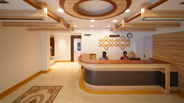 A reception area with a wooden reception desk, a clock on the wall, and a patterned rug on the floor. - Hotel Palmyra Grand Inn (Tirunelveli)