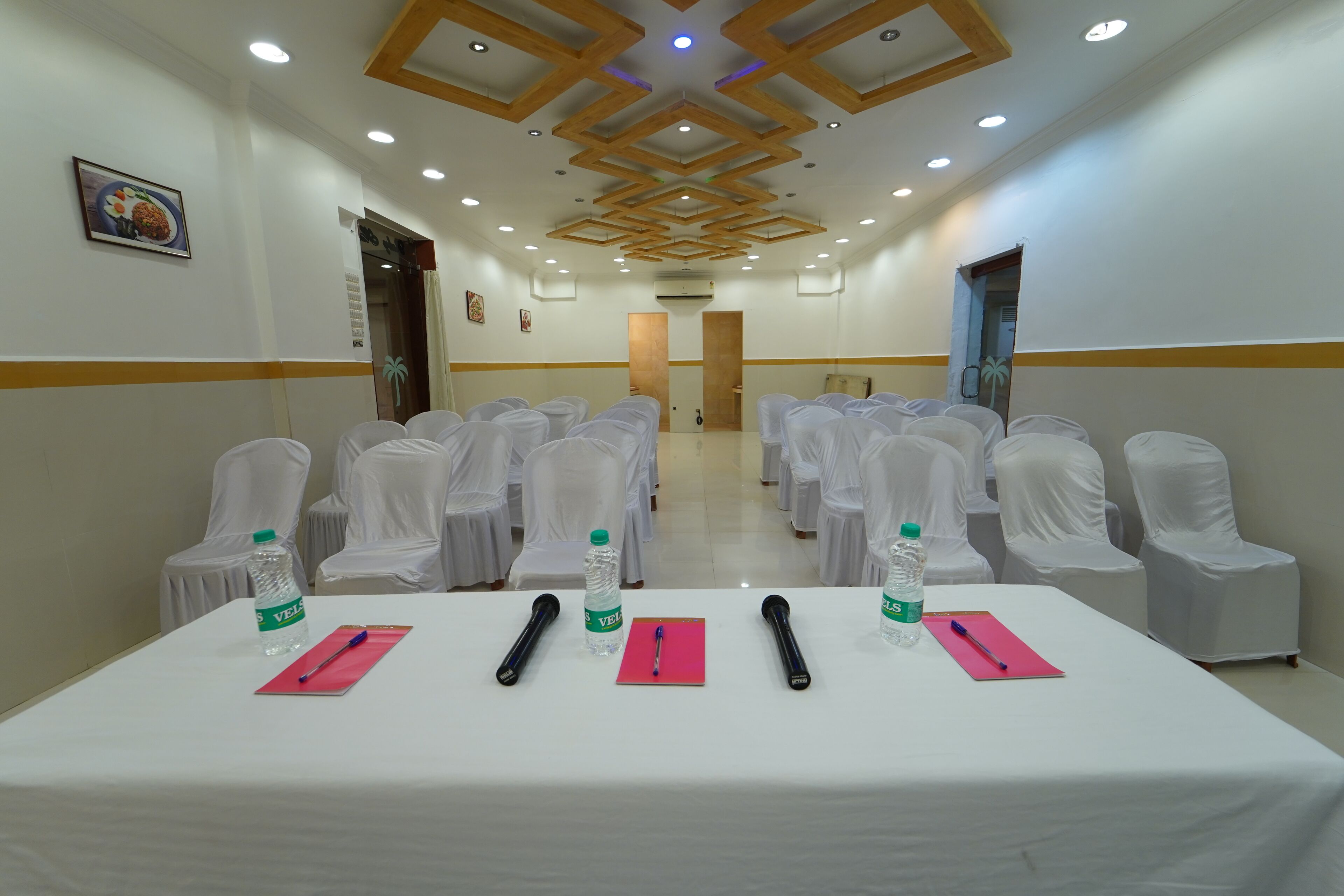 A conference room with white chairs, a table with microphones, water bottles, and a white tablecloth.