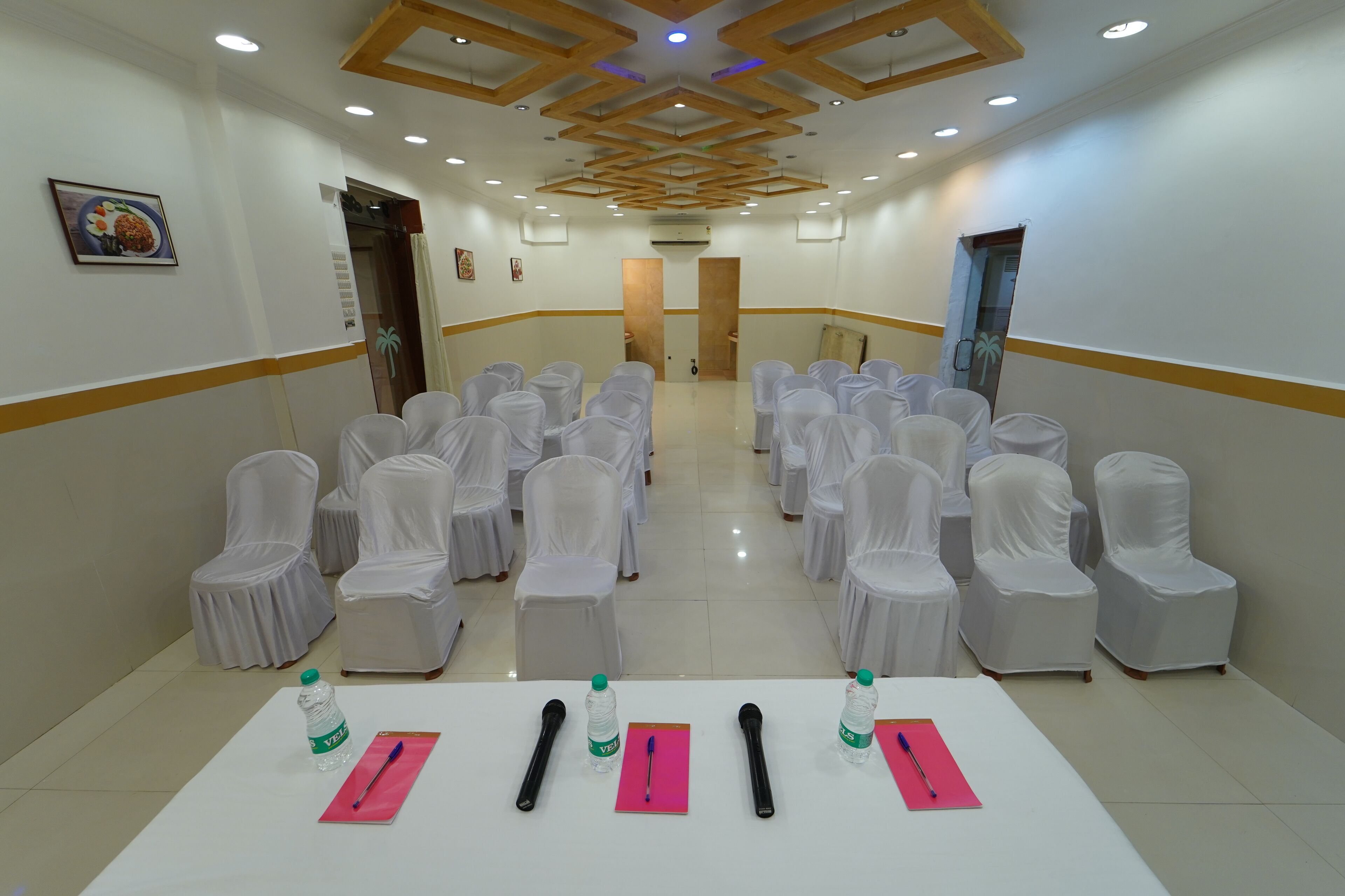 Une salle de conférence avec des rangées de chaises recouvertes de tissu blanc, une table blanche ornée de bouteilles d’eau et de stylos, et un plafond avec des poutres en bois et des luminaires encastrés.