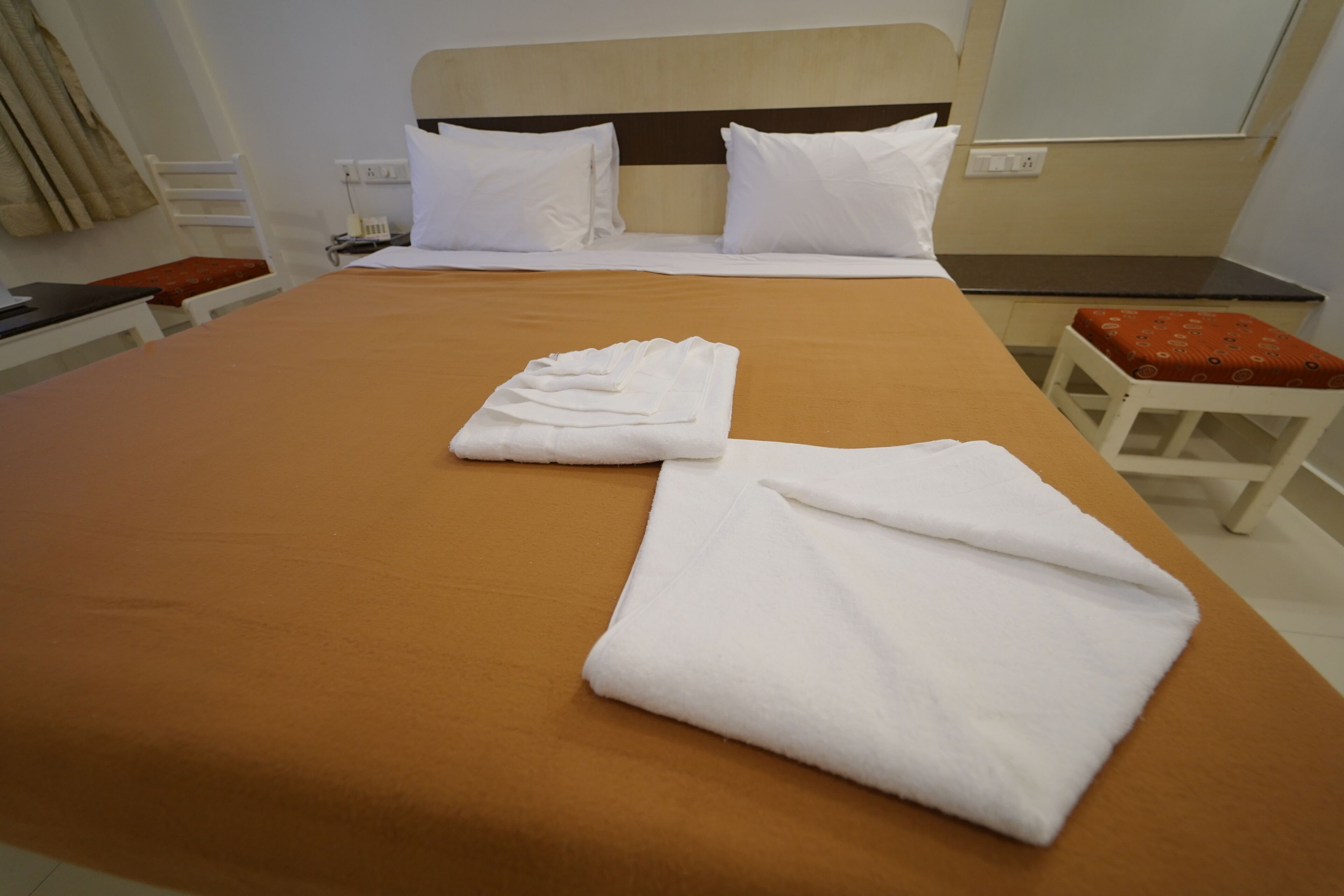 Een hotelkamer met een bed, twee witte handdoeken, een houten hoofdeinde, een nachtkastje en een rode, beklede kruk.