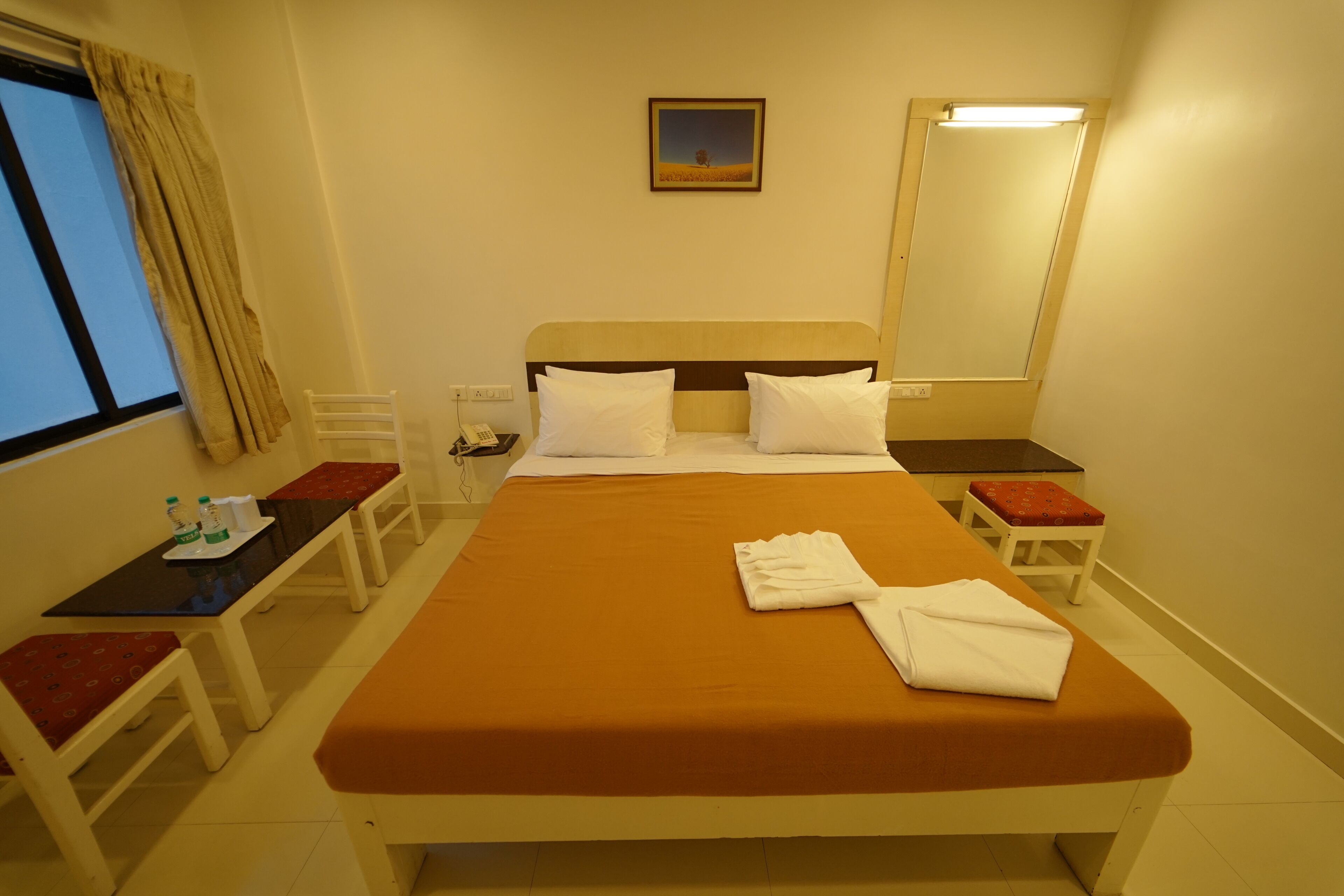 A hotel room with a bed, a desk, a chair, a small table, a mirror, and a window with curtains.