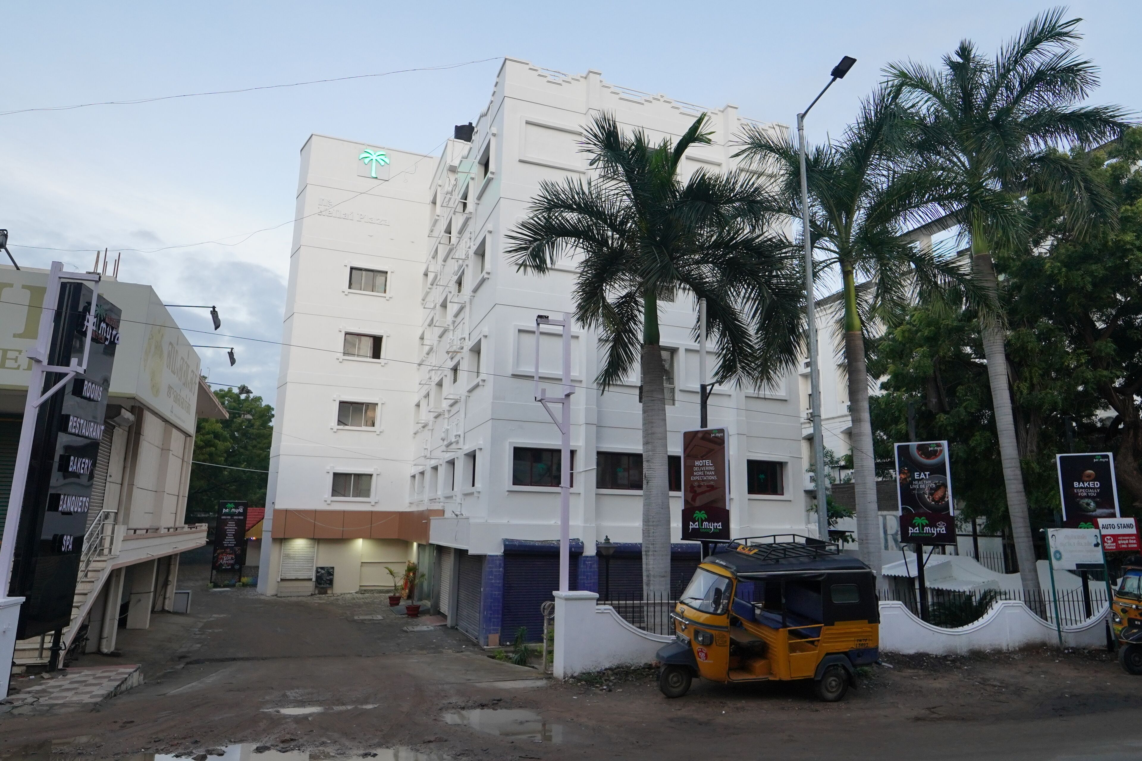 A multi-story hotel with a white facade, palm trees, and a parked auto-rickshaw.