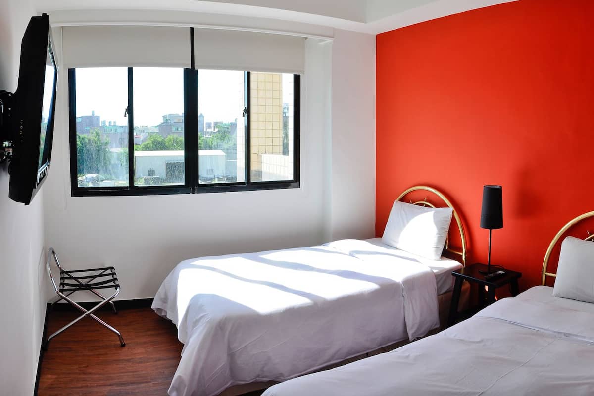 Ein Hotelzimmer mit zwei Betten, einer roten Akzentwand, einem Fenster mit Blick und einem Schreibtisch mit Telefon sowie einem Stuhl.