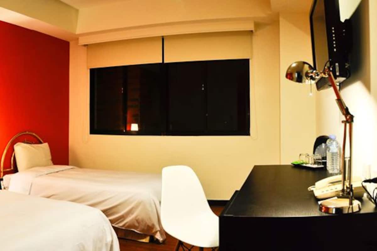 Ein Hotelzimmer mit zwei Betten, einem Schreibtisch, einem Stuhl, einer Schreibtischlampe und einem großen Fenster.