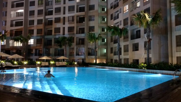 A swimming pool with a person in the water, surrounded by palm trees and lounge chairs. - Cyber Masteri Thao Dien Apartment (Ho Chi Minh City)