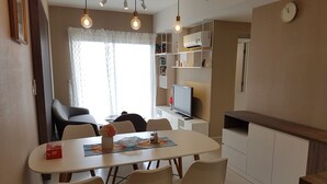 A modern dining area with a white table, four chairs, and pendant lighting. A television is mounted on the wall, and there is a clock above it. - Cyber Masteri Thao Dien Apartment (Ho Chi Minh City)
