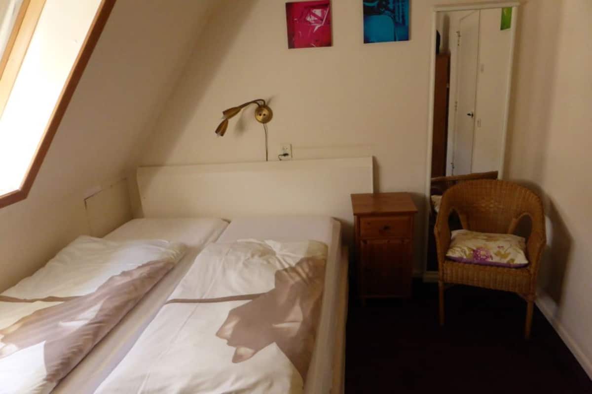Ein kleiner, schwach beleuchteter Raum mit einem Einzelbett, einem Nachttisch, einem Korbstuhl und einer Tür zu einem anderen Raum.