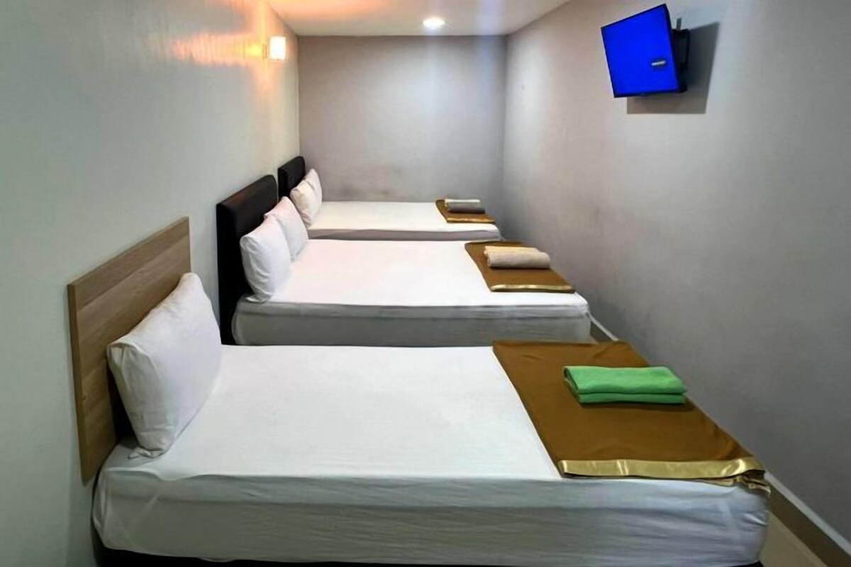 Habitación de hotel con cuatro camas individuales dispuestas en fila, cada una con cabecero de madera y ropa de cama blanca. Un televisor de pantalla plana está montado en la pared sobre las camas.