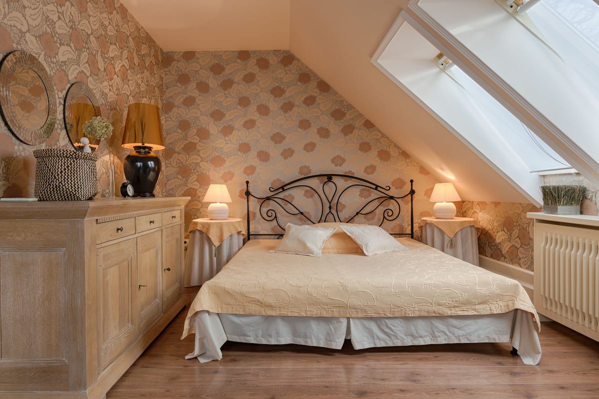 A bedroom with a sloped ceiling, a bed with a beige bedspread, two bedside lamps, a wooden dresser, and a radiator.
