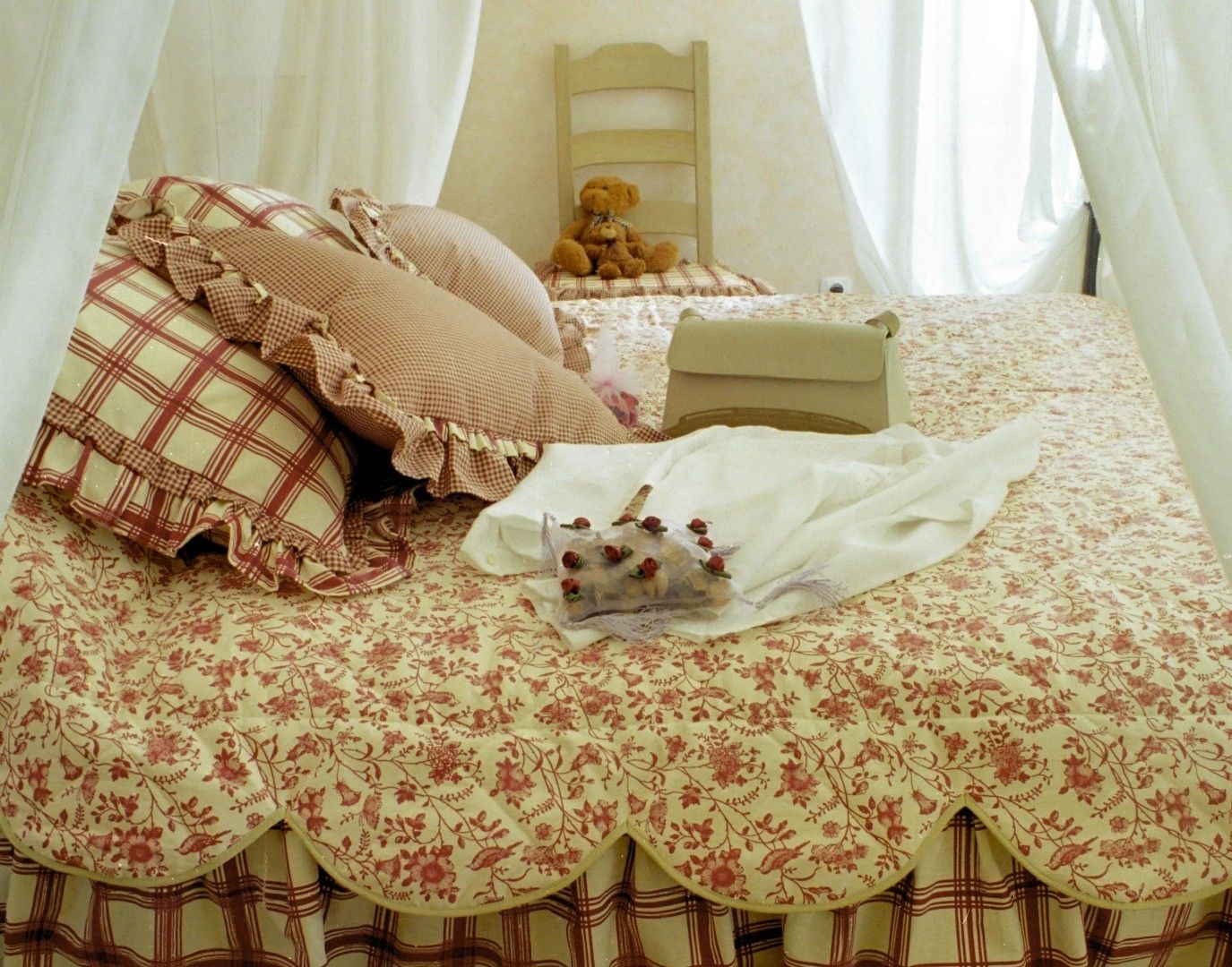 A bed with floral bedding, a checkered pillow, a teddy bear, and a bedside table with a lamp.