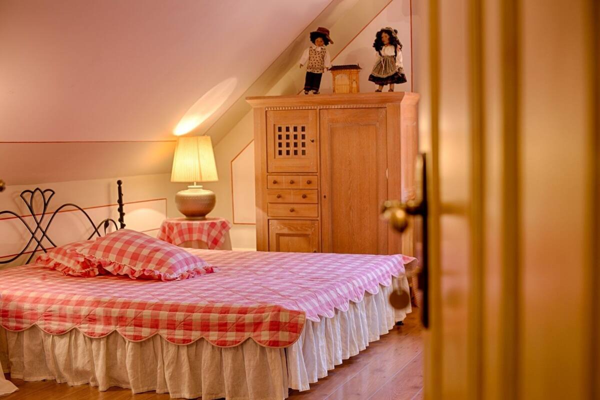 A bedroom with a checkered bedspread, a wooden wardrobe, a nightstand with a lamp, and a small table with dolls.