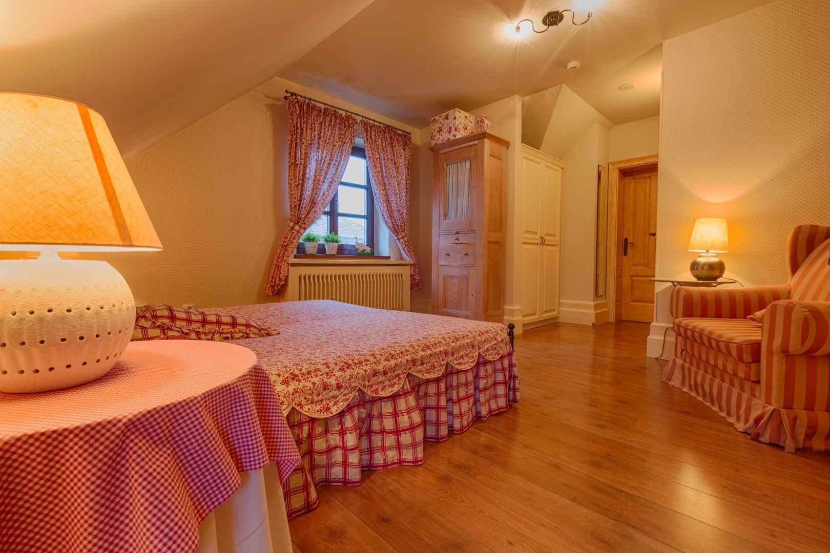 Ein gemütliches Schlafzimmer mit Bett, Nachttisch mit Lampe, Fenster mit Vorhängen, Heizkörper, Holztür und einem gestreiften Sessel.