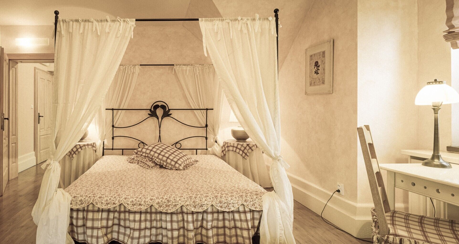 A four-poster bed with a floral bedspread and plaid skirt, a bedside table with a lamp, and a framed picture on the wall.