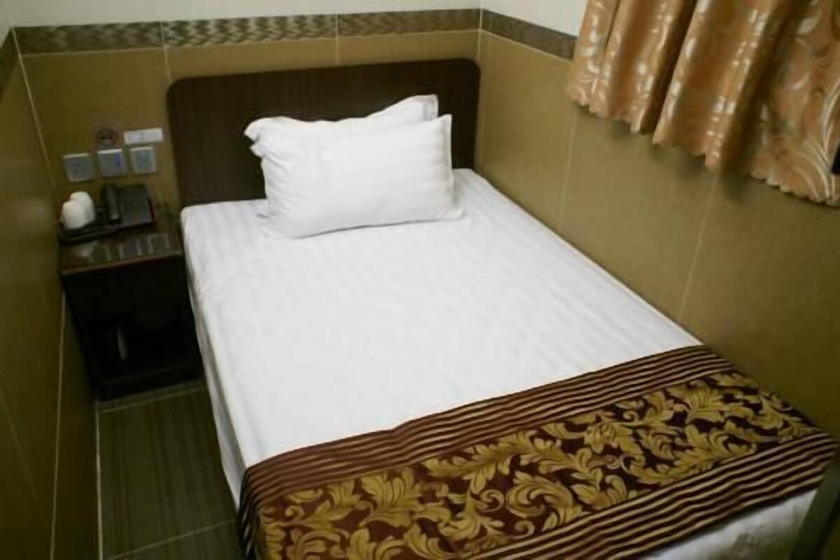 Una habitación pequeña con cama individual, colcha marrón estampada, almohada blanca y mesita de noche con lámpara y pequeña mesa.
