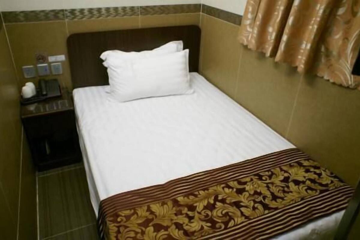 Una habitación pequeña con una cama individual, colcha marrón estampada, almohada blanca, mesita de noche con lámpara y cortina marrón.