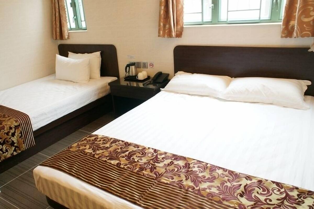 Habitación de hotel con dos camas individuales, una mesita de noche con un teléfono y una tetera, y una ventana con cortinas.