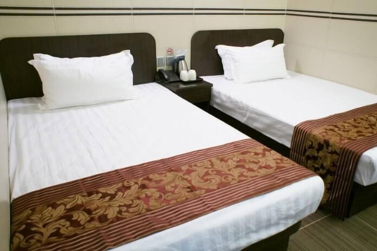 Habitación de hotel con dos camas individuales, cada una con ropa blanca y cabeceros marrones. Hay una mesita de noche pequeña con una lámpara y un teléfono.