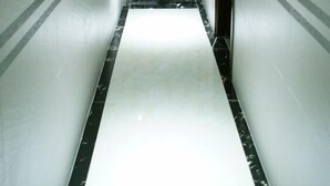 Un pasillo estrecho y bien iluminado, con piso blanco y detalles de mármol negro, que conduce a una puerta de salida con luz azul.